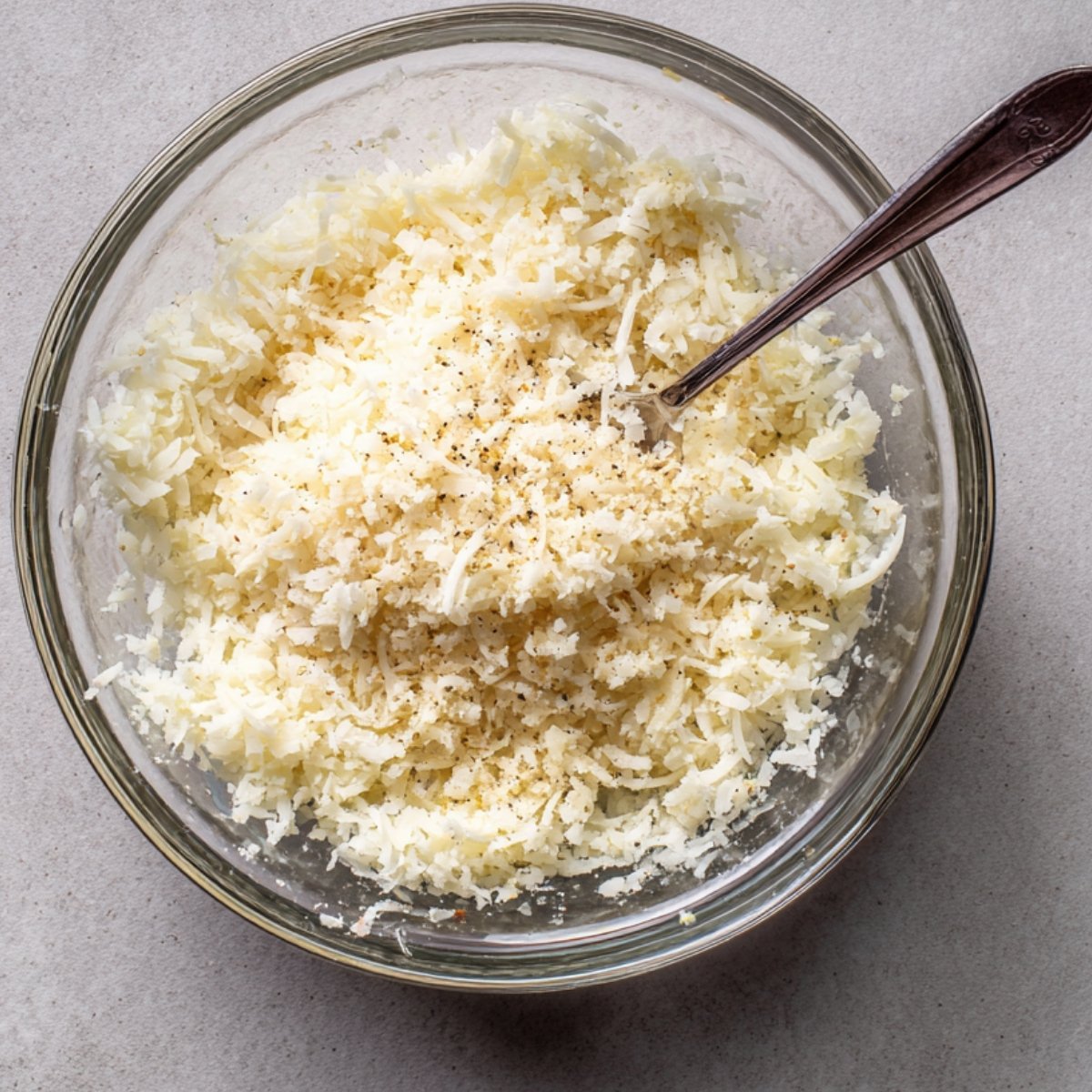 Glass bowl filled with grated Parmesan cheese, seasoned with black pepper, with a spoon inside.