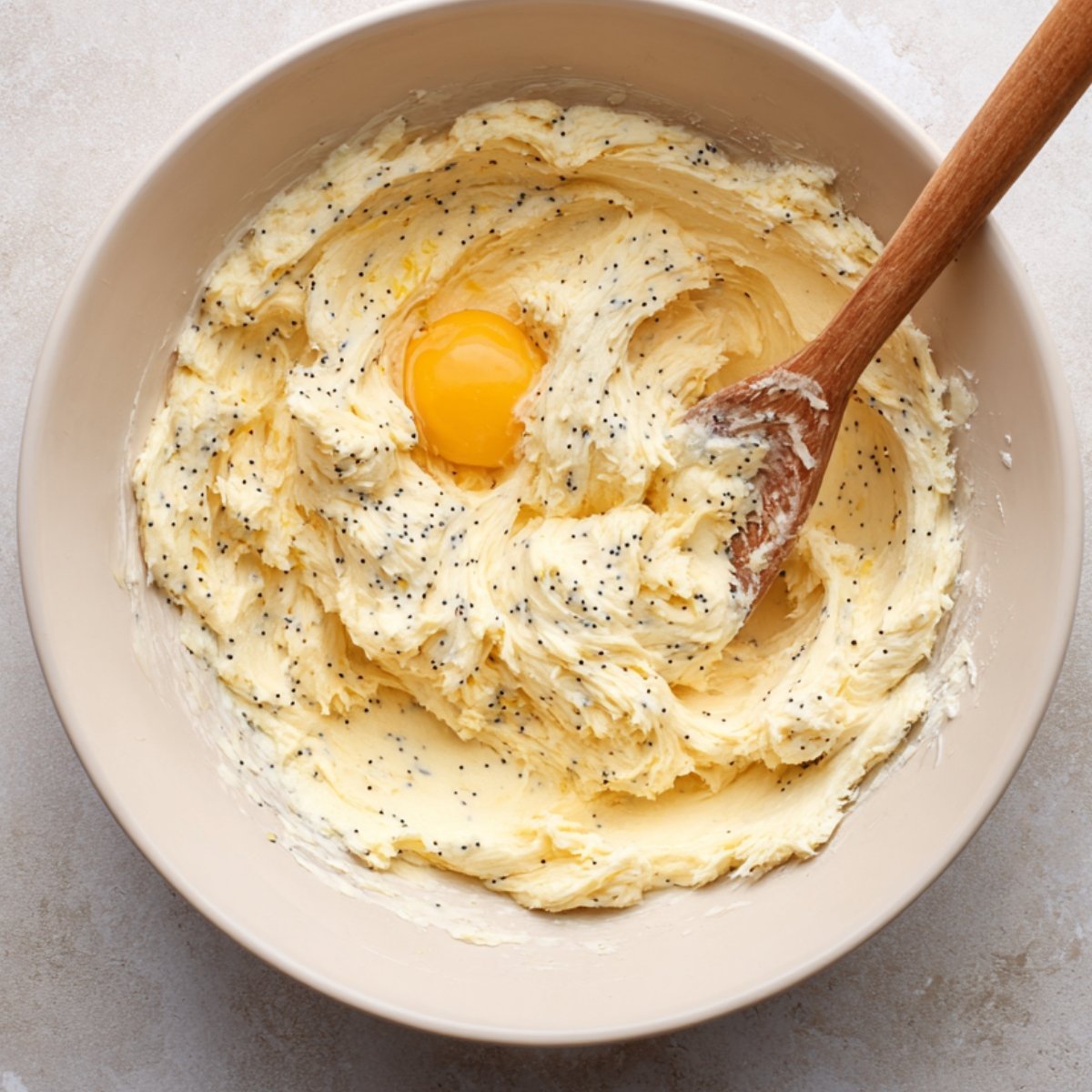 Creamy butter mixture with poppy seeds and an egg yolk, being mixed with a wooden spoon.