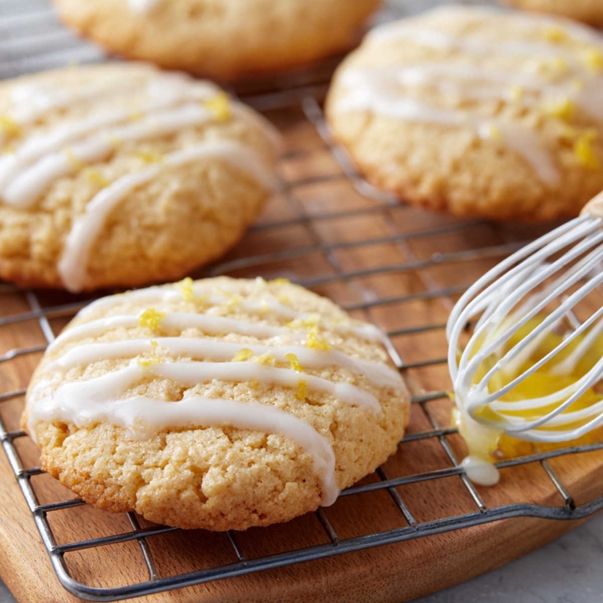 Golden cookies drizzled with lemon glaze cooling on a wire rack, with a whisk holding glaze nearby.
