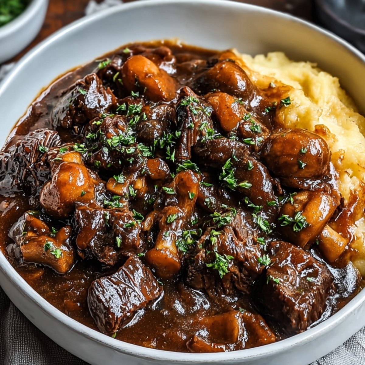 A bowl of hearty Beef Bourguignon recipe served with mashed potatoes, with tender beef chunks and carrots in a thick, dark gravy, topped with fresh parsley.