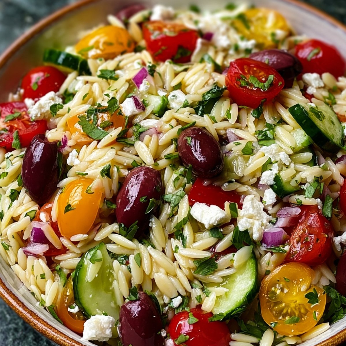 Close-up of a vibrant Greek orzo salad with red and yellow cherry tomatoes, cucumbers, Kalamata olives, fresh parsley, and crumbled feta cheese in a serving bowl.