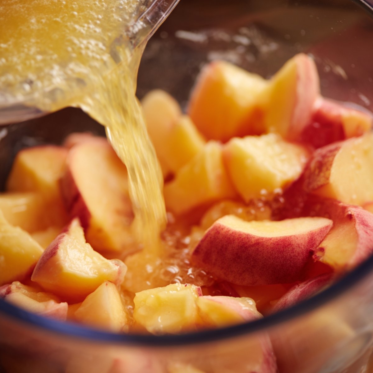 Chunks of fresh peaches in a blender with lemon juice being poured over them before blending.
