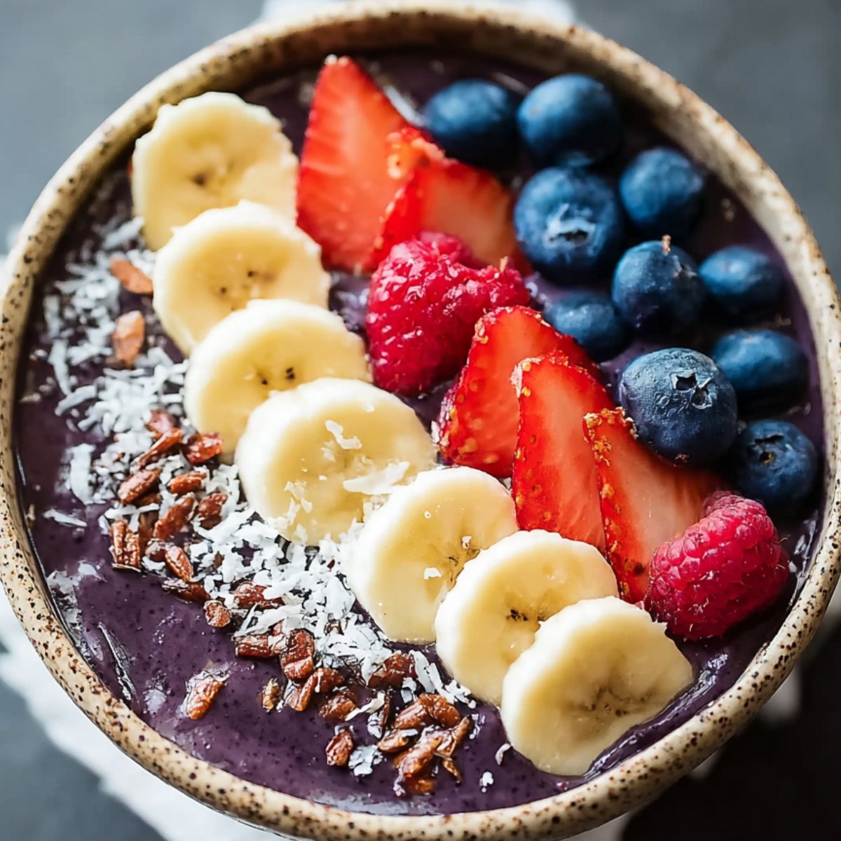 A finished acai bowl recipe topped with banana slices, strawberries, raspberries, blueberries, shredded coconut, and crunchy granola.