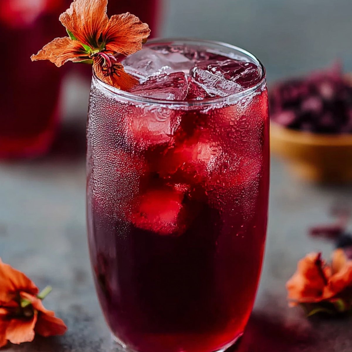 A chilled glass of deep red iced hibiscus tea recipe filled with ice cubes, garnished with an orange hibiscus flower on the rim.