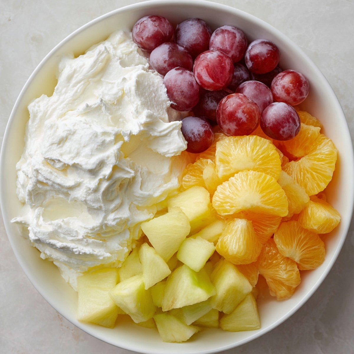 A white bowl filled with whipped cream, red grapes, orange segments, pineapple chunks, and honeydew melon pieces.