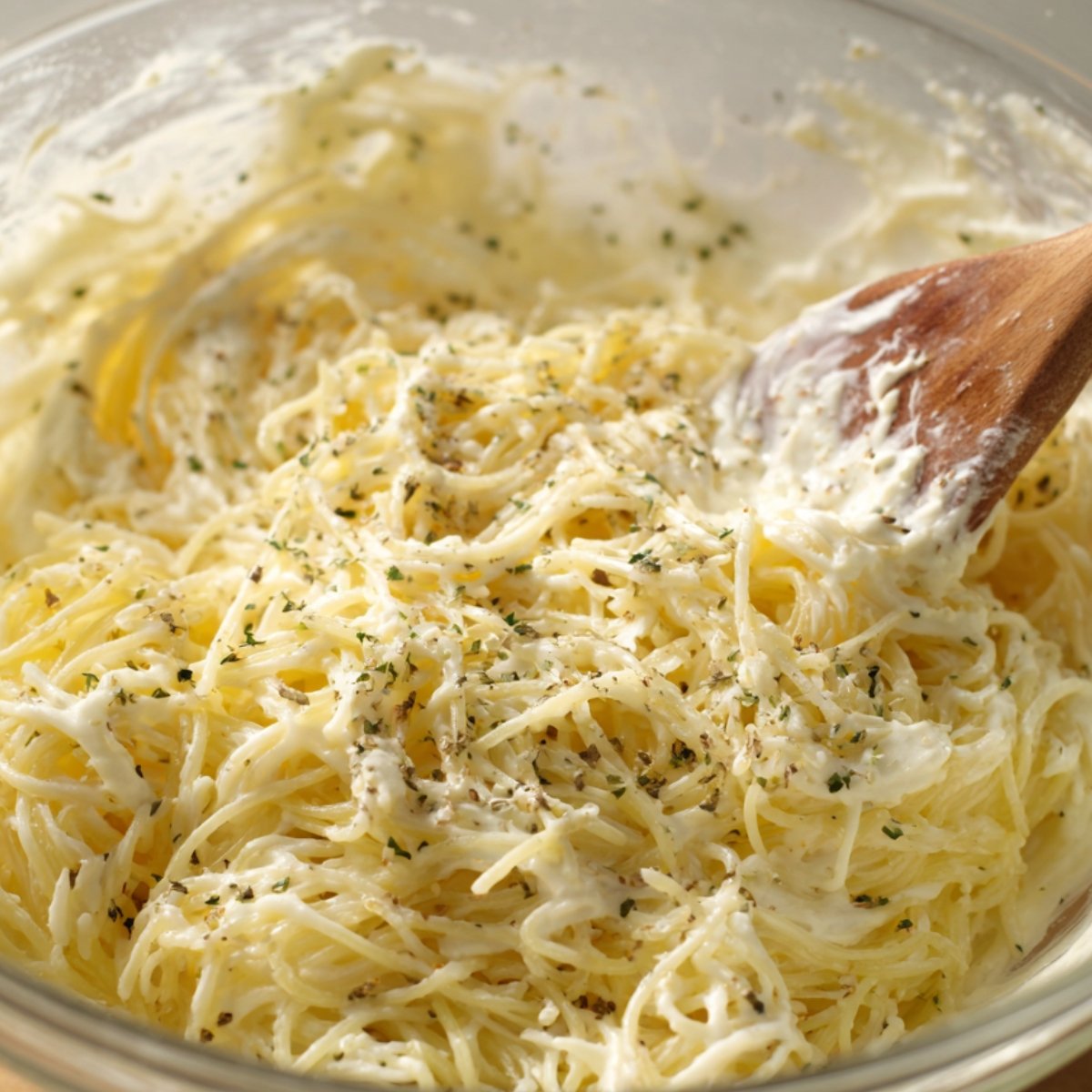 "Spaghetti mixed with cream cheese and herbs, being stirred with a wooden spoon in a glass bowl."