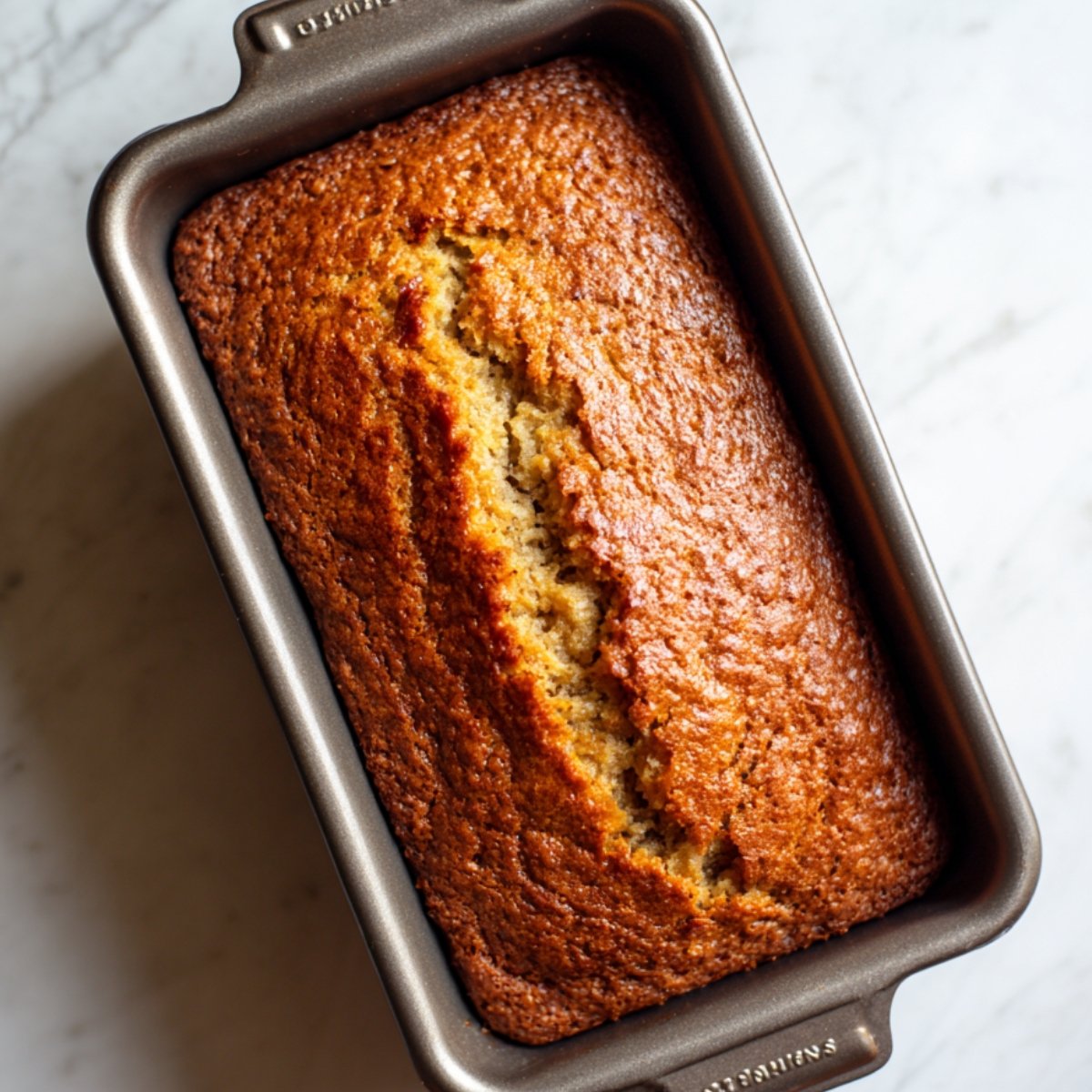 A freshly baked golden-brown loaf of banana bread in a rectangular metal pan, with a split down the center showing its moist texture.