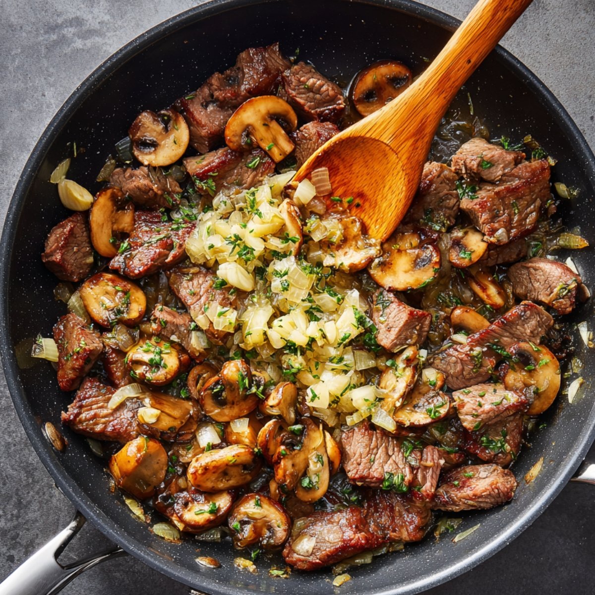 "Beef, mushrooms, and onions sizzling in a skillet, with herbs and seasonings sprinkled on top."