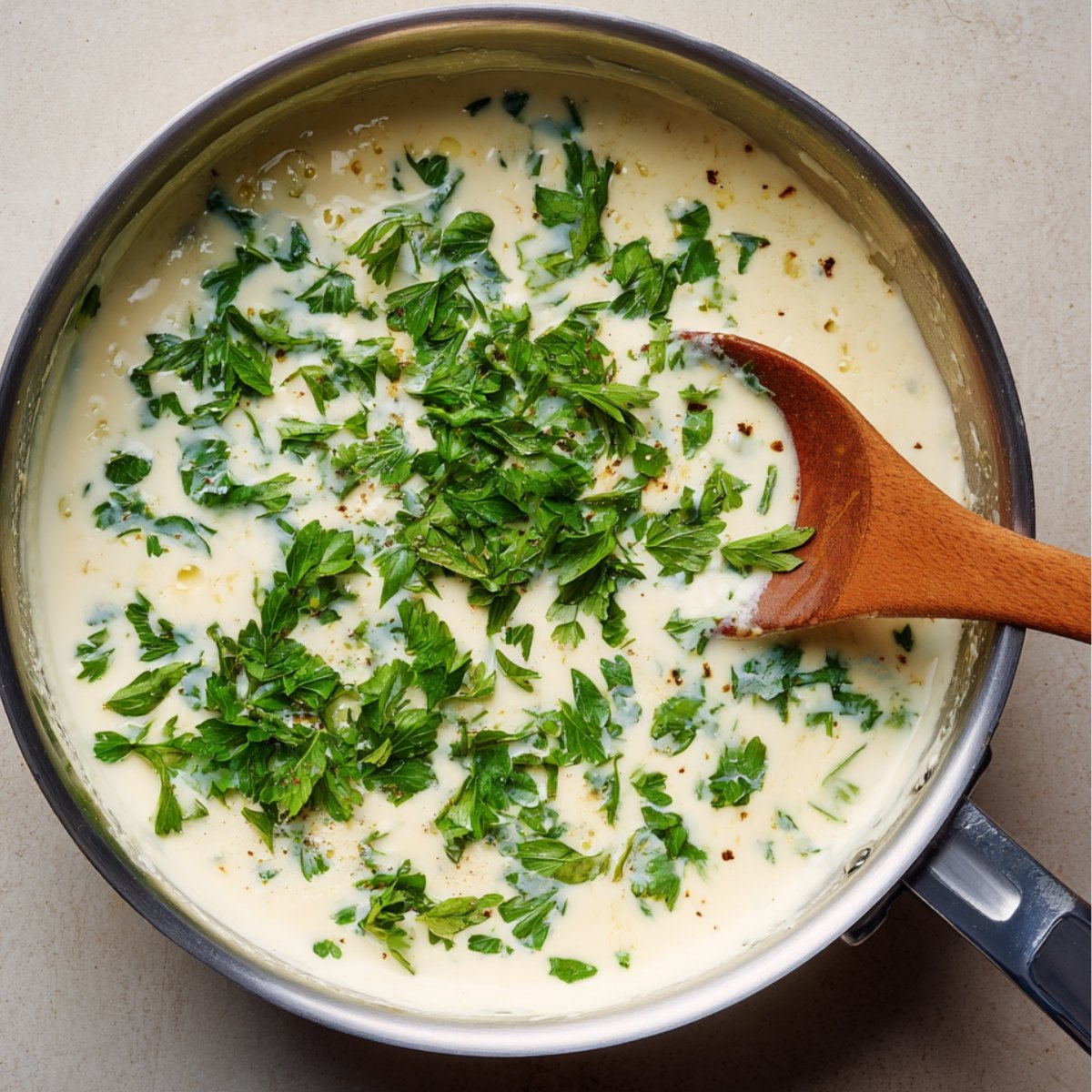 "A creamy sauce with fresh parsley sprinkled on top, stirred with a wooden spoon in a pan."