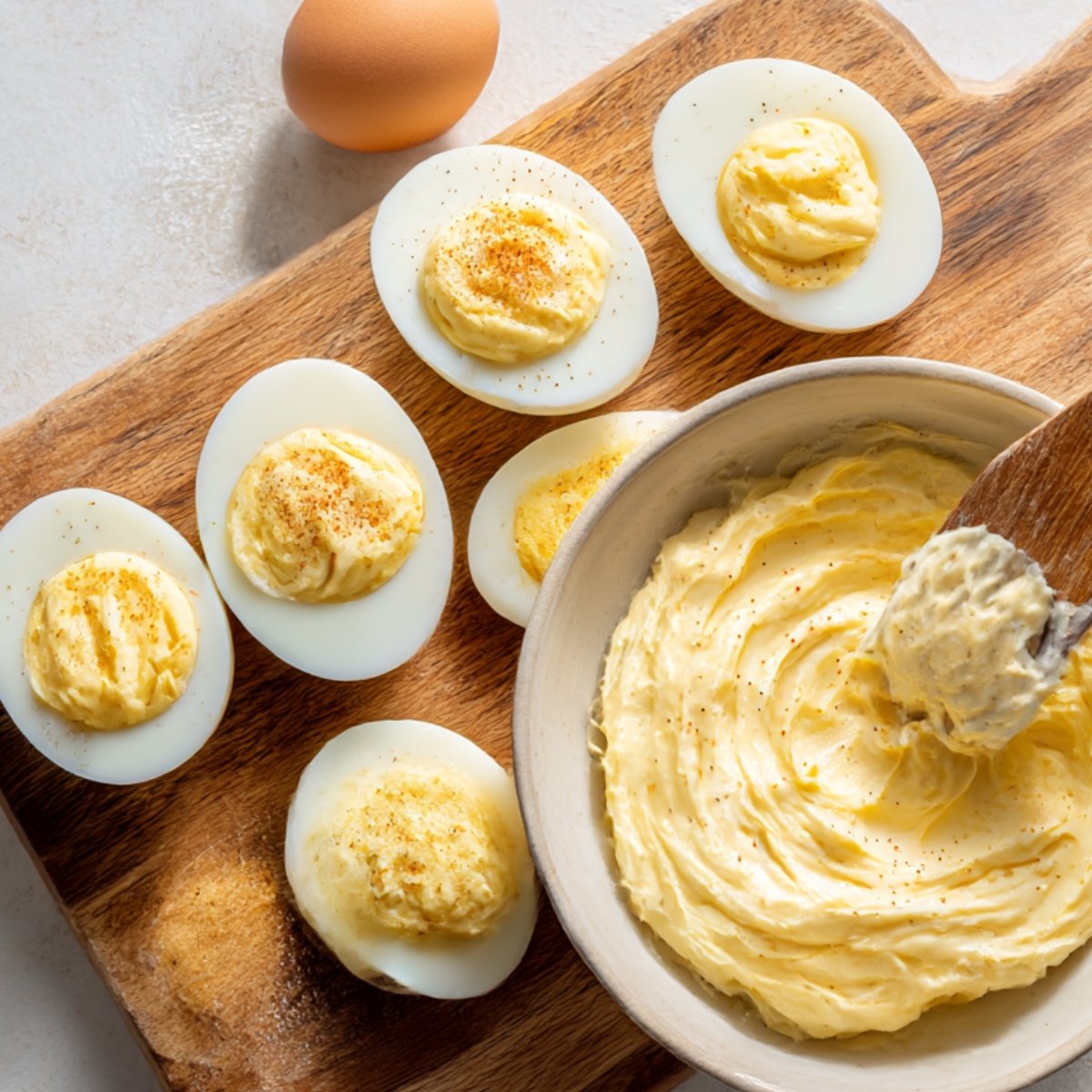 Halved hard-boiled eggs filled with creamy yolk mixture on a wooden board, with a bowl of the prepared filling and an egg in the background.