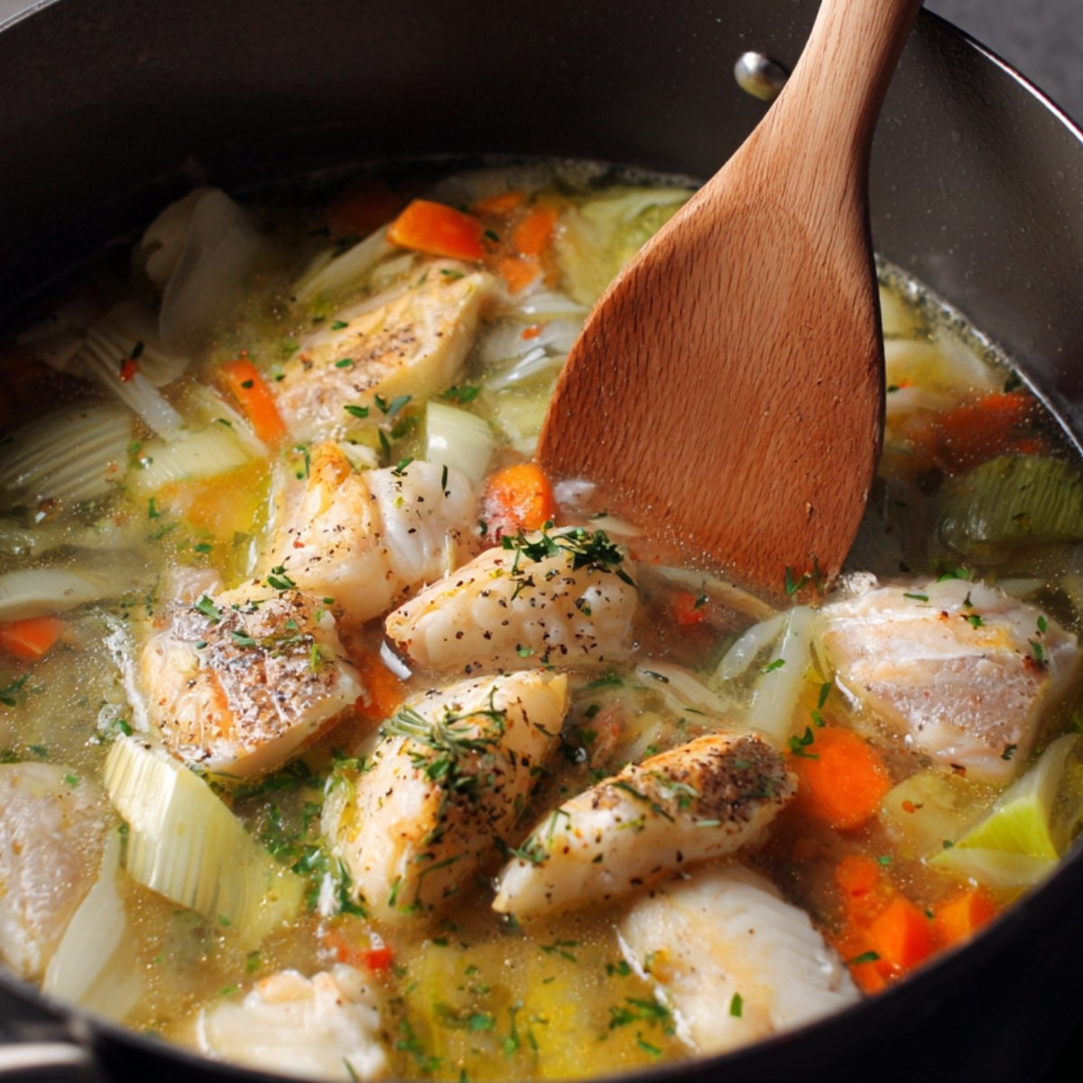 A simmering pot of fish soup with chunks of white fish, carrots, leeks, and herbs in a clear broth.