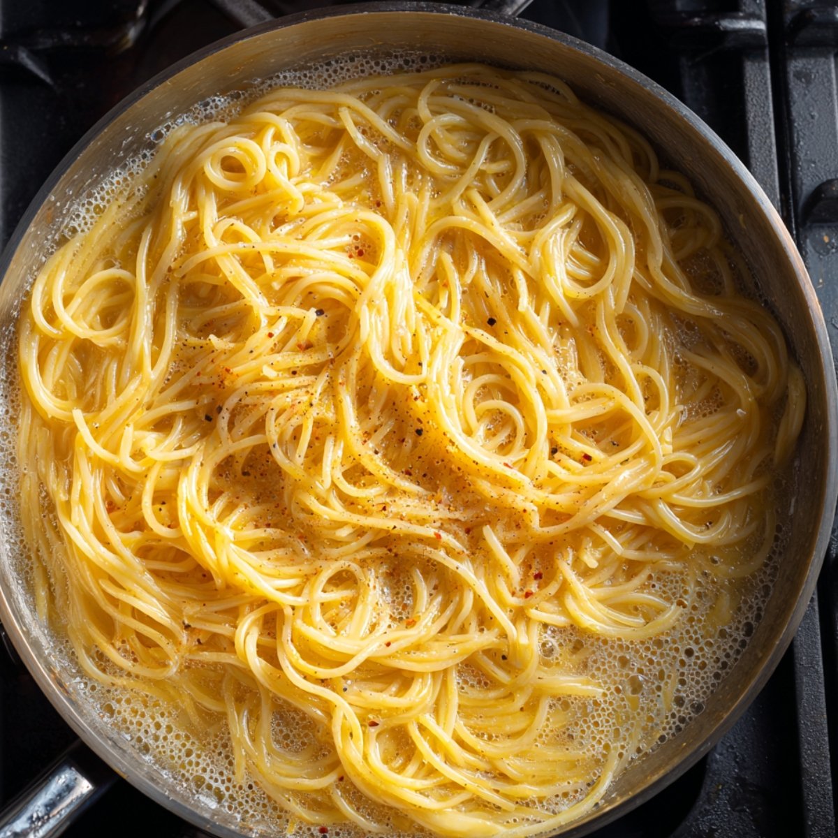 Spaghetti coated in a rich, golden sauce cooking in a pan, seasoned lightly with black pepper.