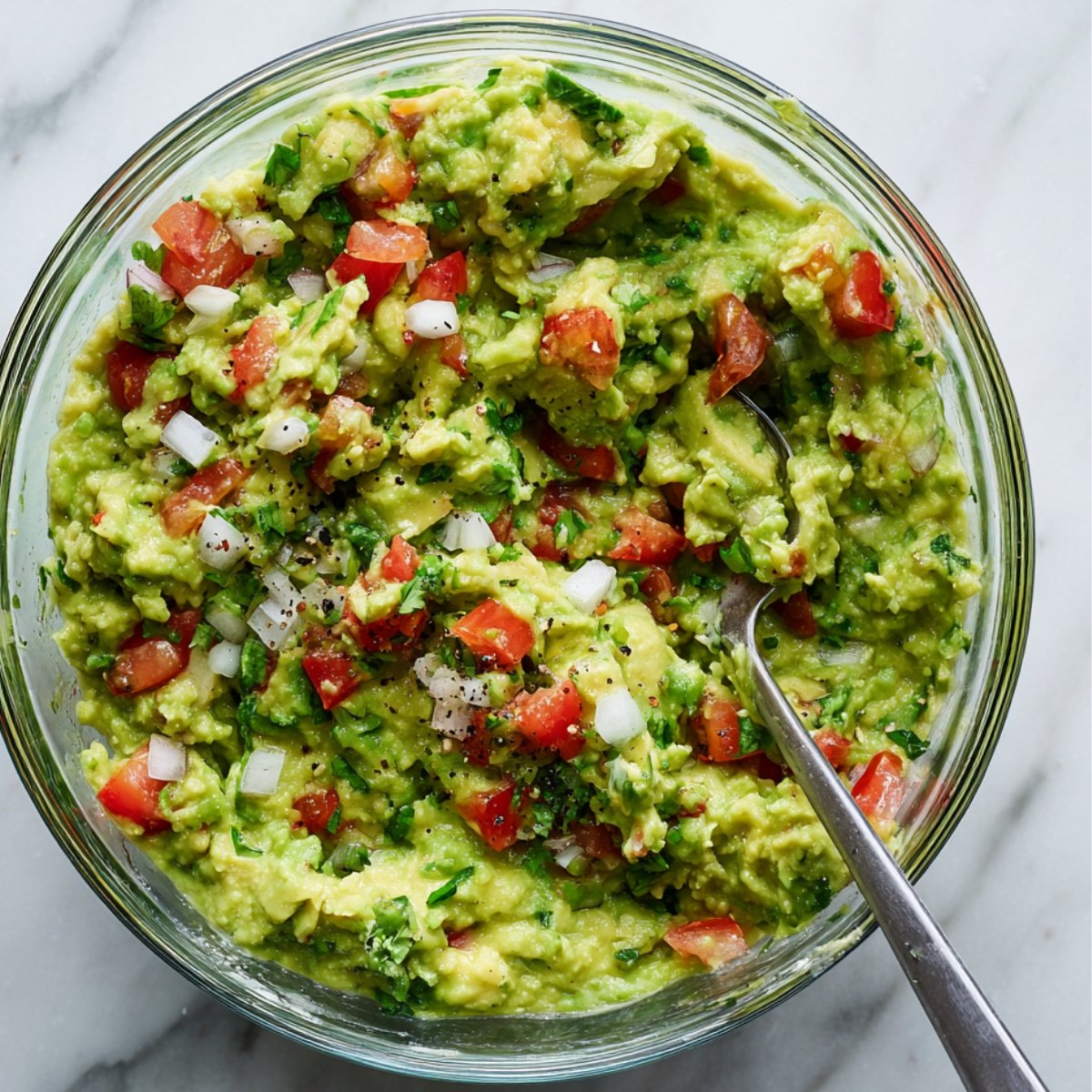 A glass bowl filled with chunky guacamole mixed with diced tomatoes, onions, cilantro, and black pepper.
