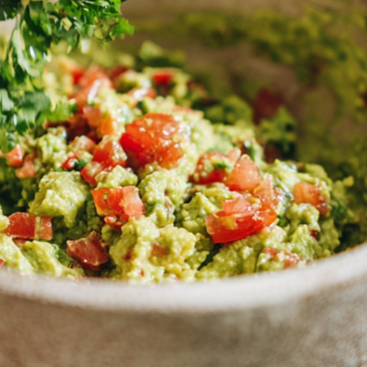 A rustic bowl of guacamole topped with fresh tomato chunks and being garnished with fresh cilantro.