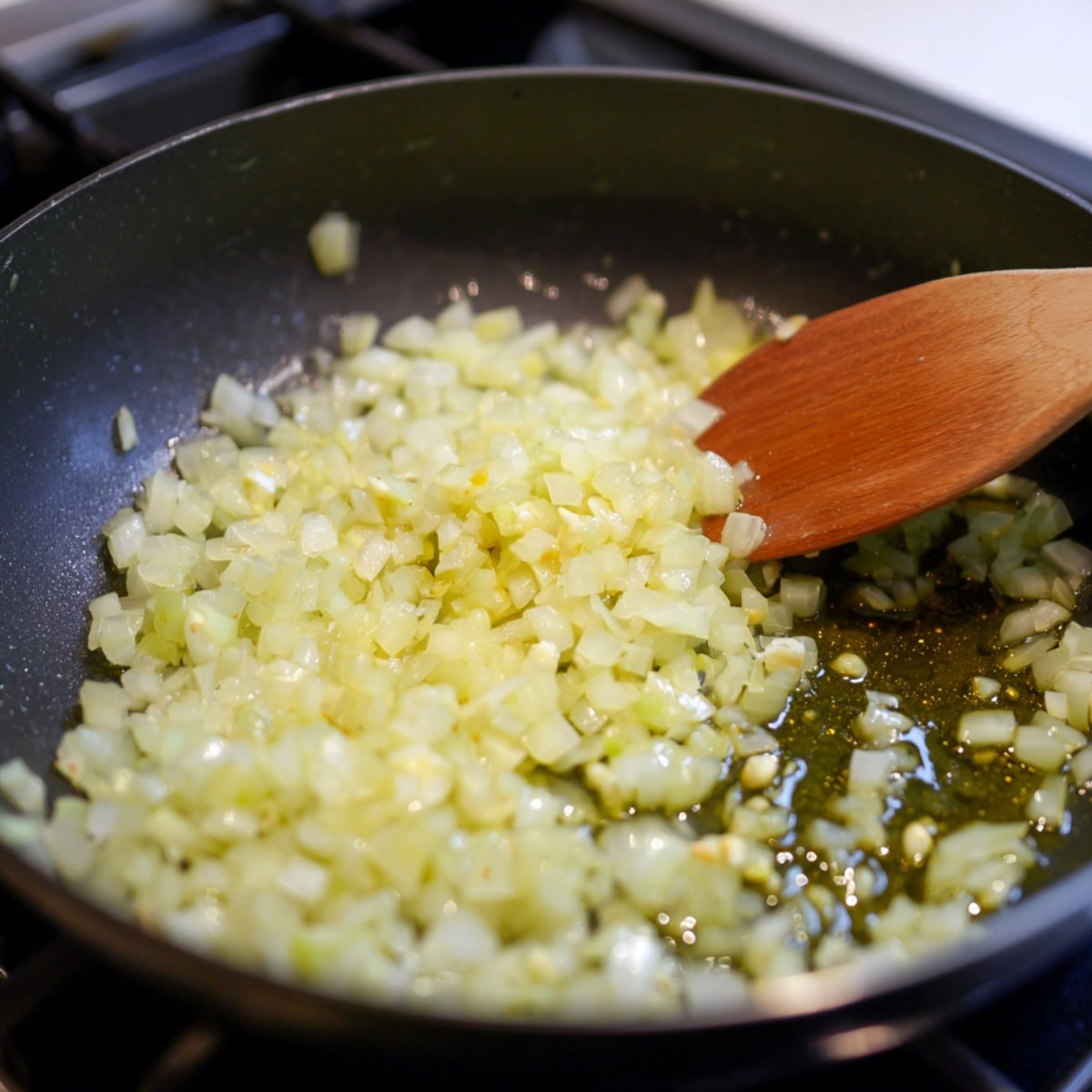 A pan of diced onions being sautéed in olive oil with a wooden spoon.