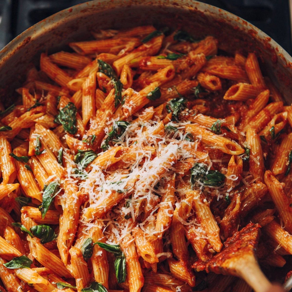 A pan filled with penne pasta tossed in a rich tomato-based sauce with fresh basil leaves, topped with grated Parmesan cheese.