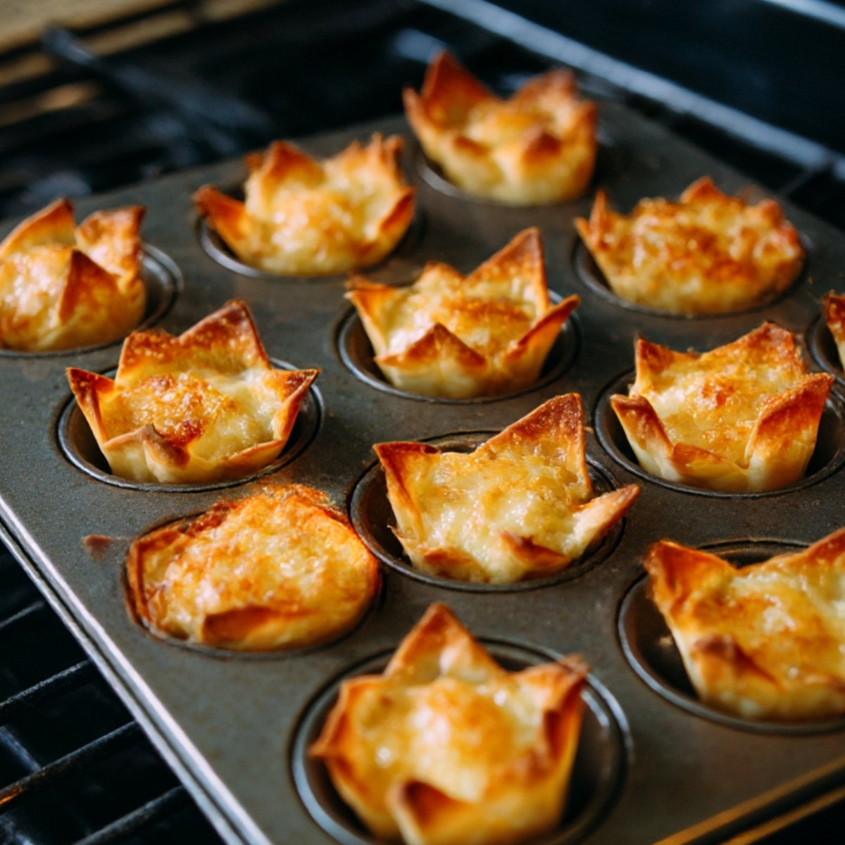 A close-up of baked taco cupcakes in a muffin tin, made with crispy wonton wrappers filled with melted cheese and golden brown edges.
