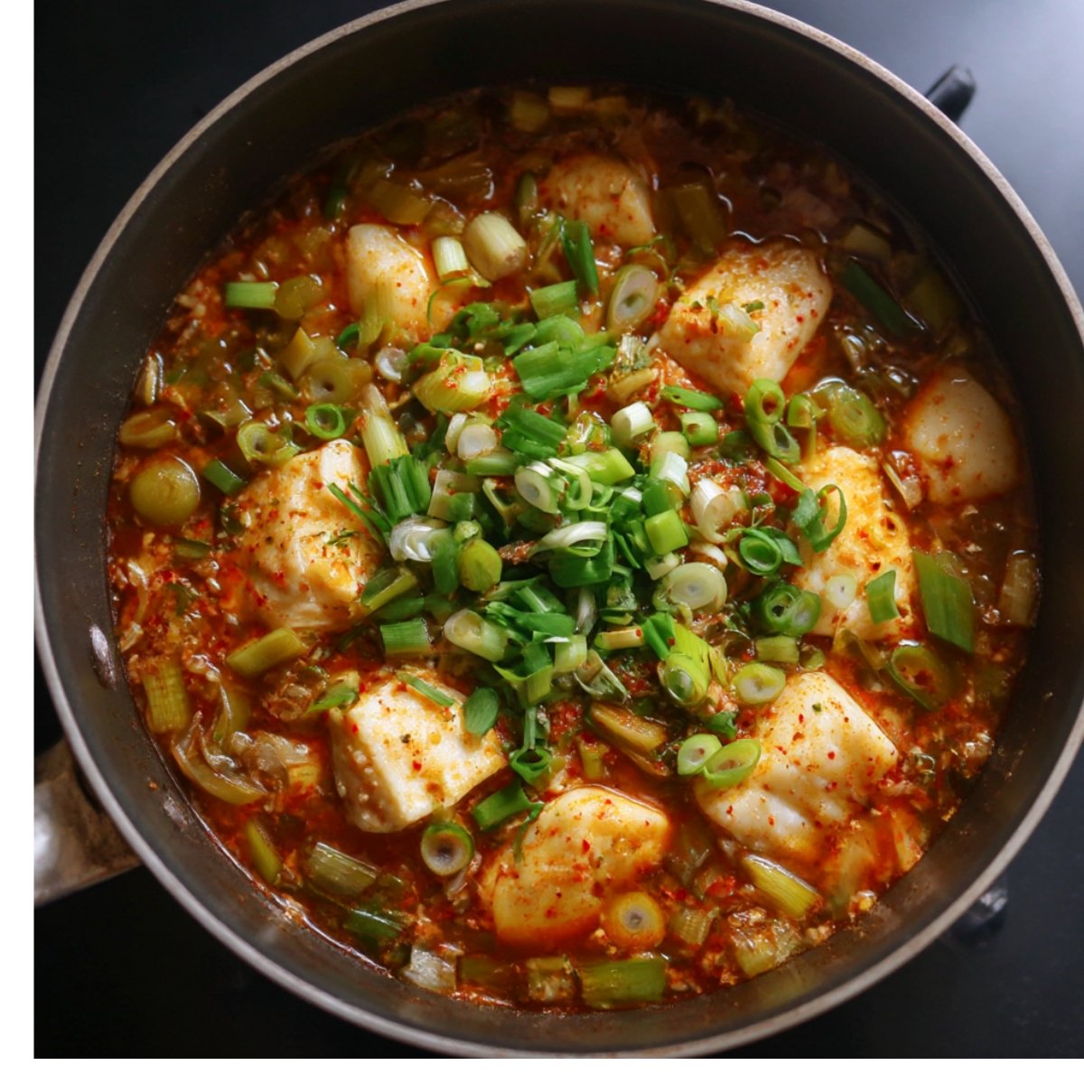A pot with boiling Tteokbokki sauce and rice cakes, topped with chopped green onions. The dish is in a simmering stage, with the sauce rich and spicy, and the rice cakes absorbing the flavors. The background is dark, showing a cozy kitchen atmosphere.