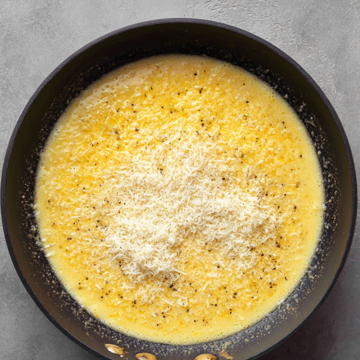 A skillet of melted Alfredo sauce base with grated Parmesan cheese sprinkled on top, lightly speckled with black pepper.