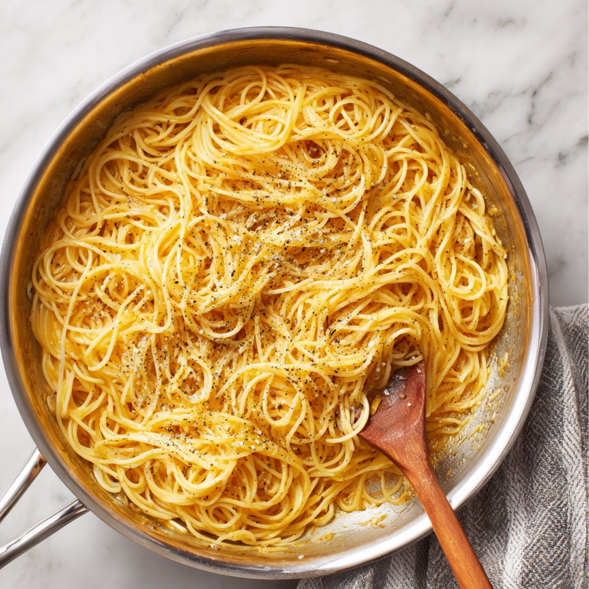 A stainless steel pan filled with spaghetti coated in creamy Alfredo sauce, topped with cracked black pepper, with a wooden spoon resting inside.