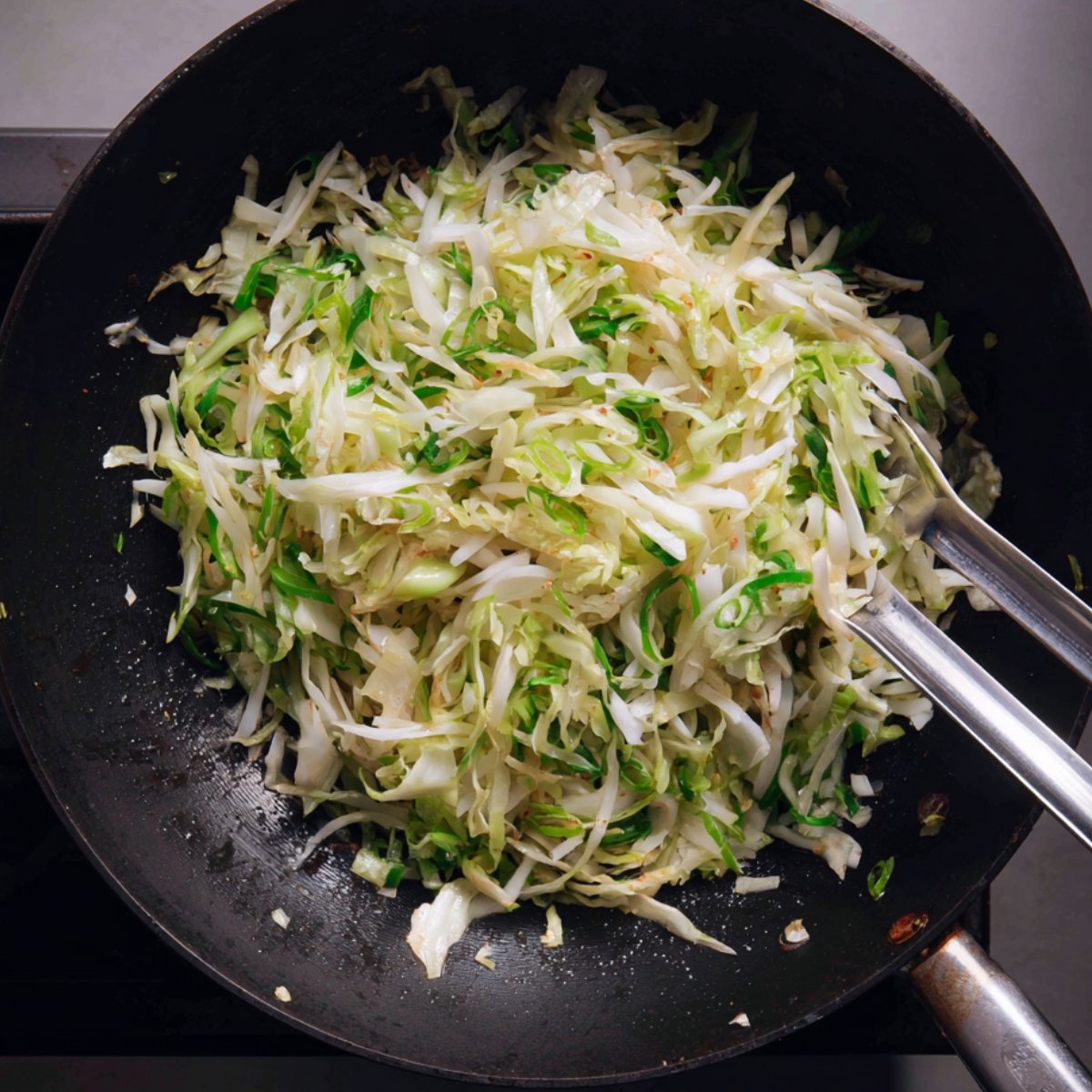 Stir-fried cabbage with some chopped green onions in a black pan with tongs.