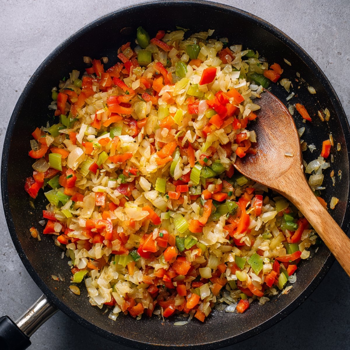 A pan of sautéed onions, red bell peppers, and green bell peppers, with a wooden spoon stirring the colorful mixture.