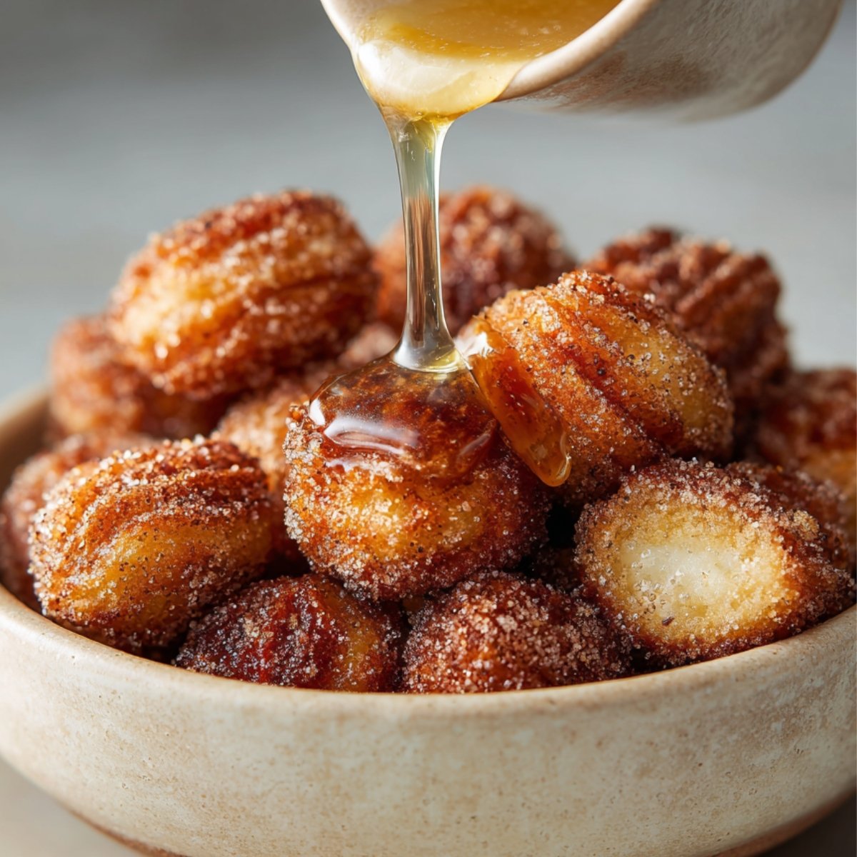 Golden churro bites in a ceramic bowl being drizzled with honey syrup, coated in cinnamon sugar.