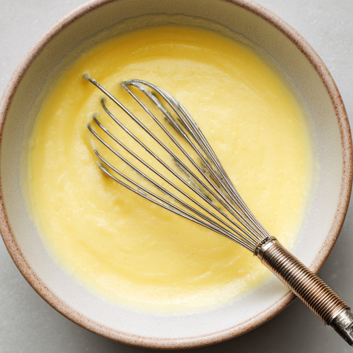 A bowl of creamy yellow mixture, possibly custard or batter, being whisked with a metal whisk in a ceramic bowl on a light surface.