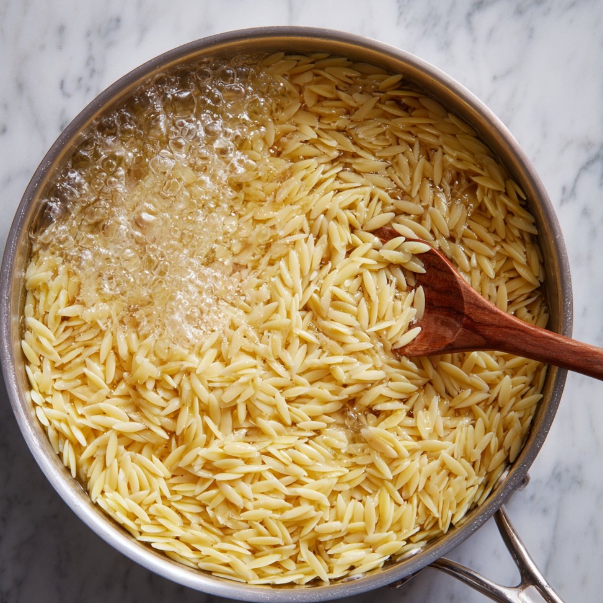 A saucepan of orzo pasta boiling in salted water with a wooden spoon stirring the pasta.