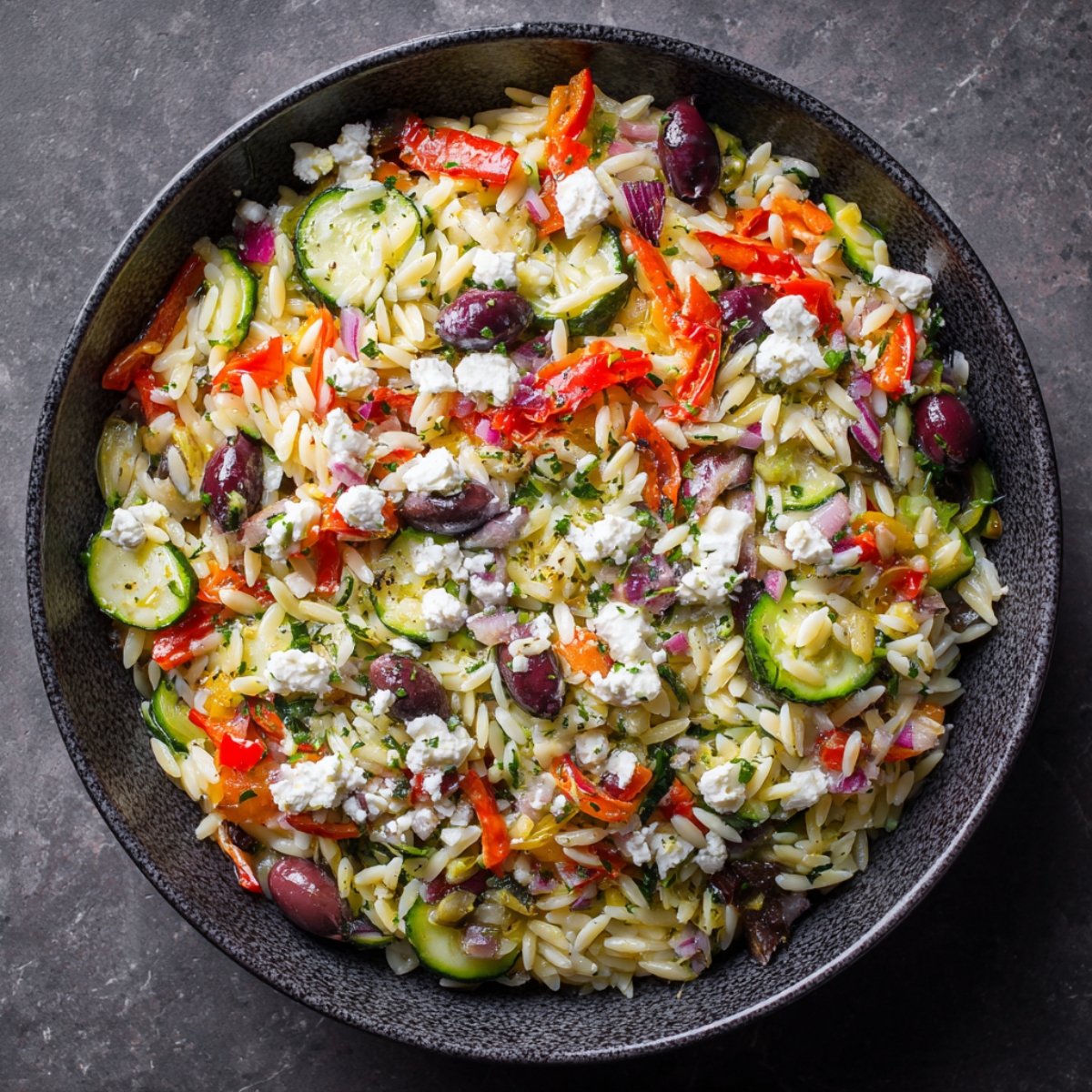 A large black bowl filled with colorful Greek orzo salad made with orzo pasta, Kalamata olives, cucumbers, cherry tomatoes, red onion, feta cheese, and fresh herbs.