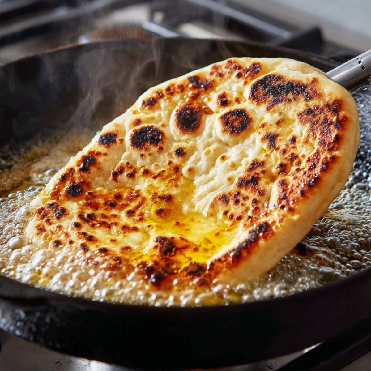 A naan bread sizzling in a hot cast iron pan with bubbling oil, golden brown spots forming on the surface.