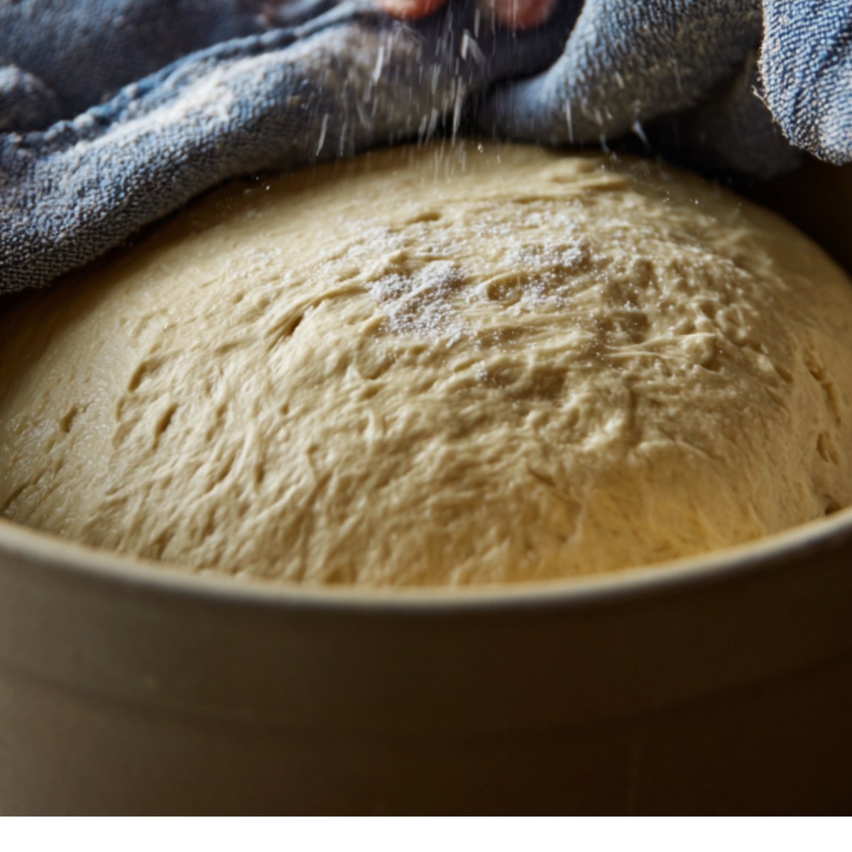 Risen dough in a bowl, soft and airy, covered partially with a cloth, sprinkled lightly with flour.
