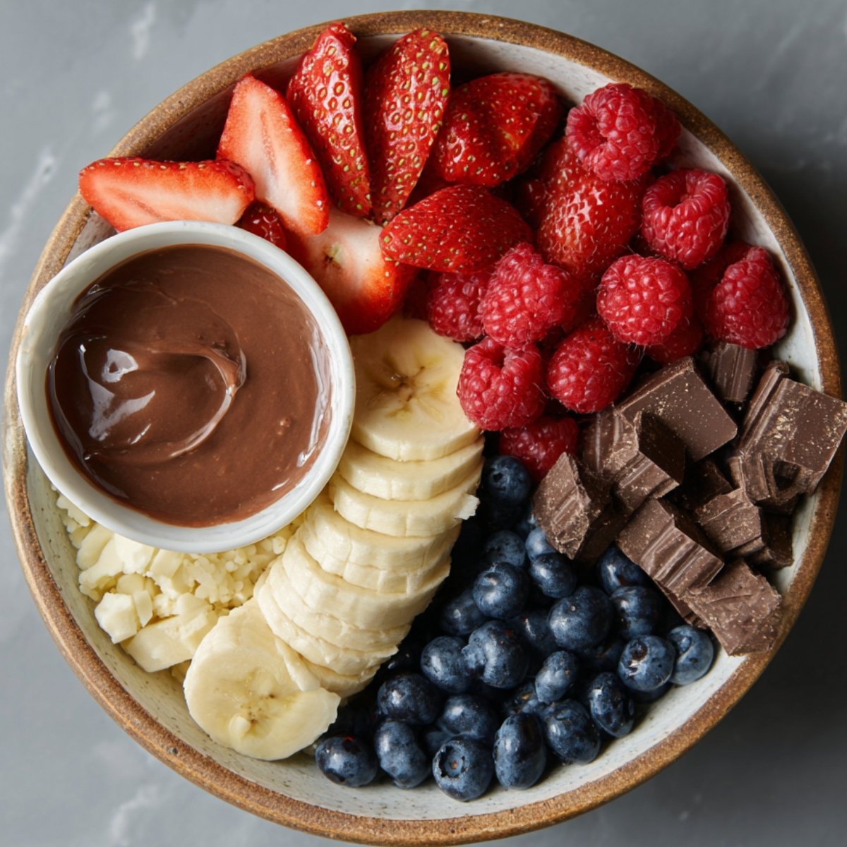 A smoothie bowl topped with fresh strawberries, raspberries, banana slices, blueberries, chunks of chocolate, and a small bowl of chocolate sauce.