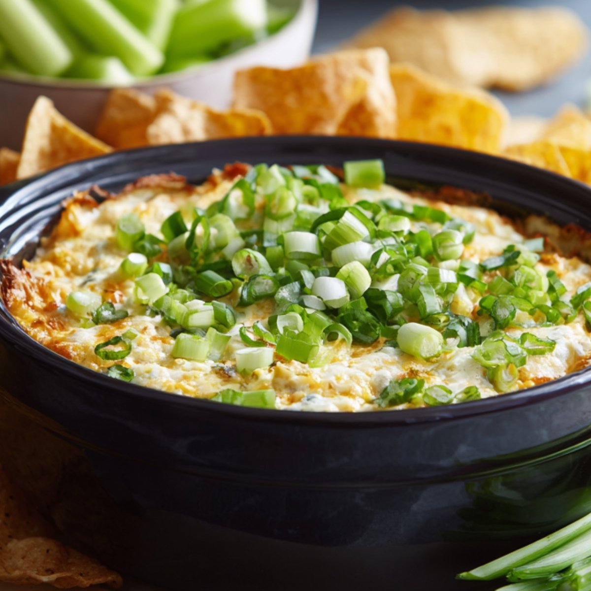 A bowl of creamy buffalo chicken dip topped with chopped green onions, surrounded by tortilla chips and celery sticks for dipping.