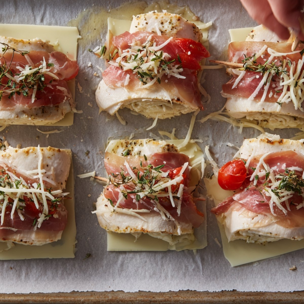 Raw chicken rollatini wrapped with prosciutto, sprinkled with shredded cheese and herbs, ready to bake.