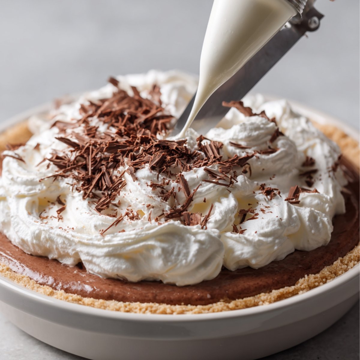 A chocolate pie topped with fluffy whipped cream and decorated with fresh chocolate shavings being piped and garnished before serving.