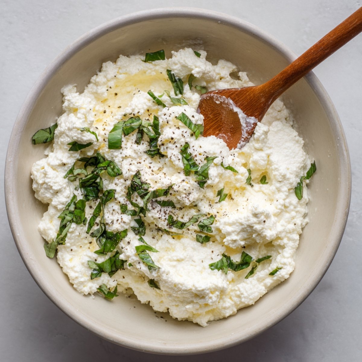 A bowl filled with ricotta cheese mixture, garnished with chopped fresh basil and cracked black pepper, with a wooden spoon resting inside.