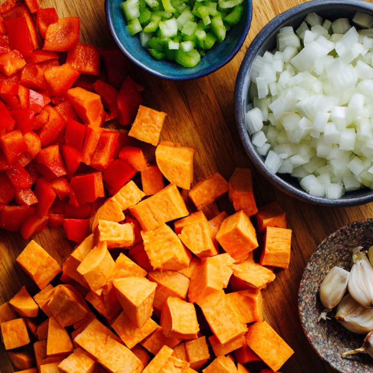 Chopped ingredients for sweet potato hash: sweet potatoes, red bell pepper, celery, onion, and garlic cloves on a wooden board.
