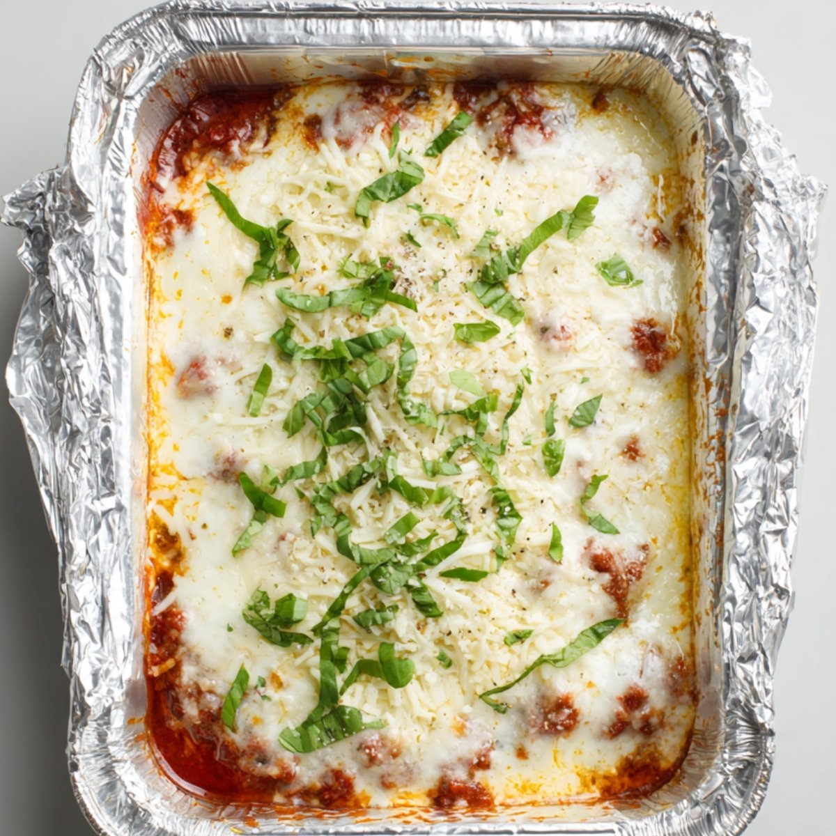 "Freshly baked zucchini lasagna topped with melted mozzarella cheese and garnished with chopped basil in a foil baking dish."