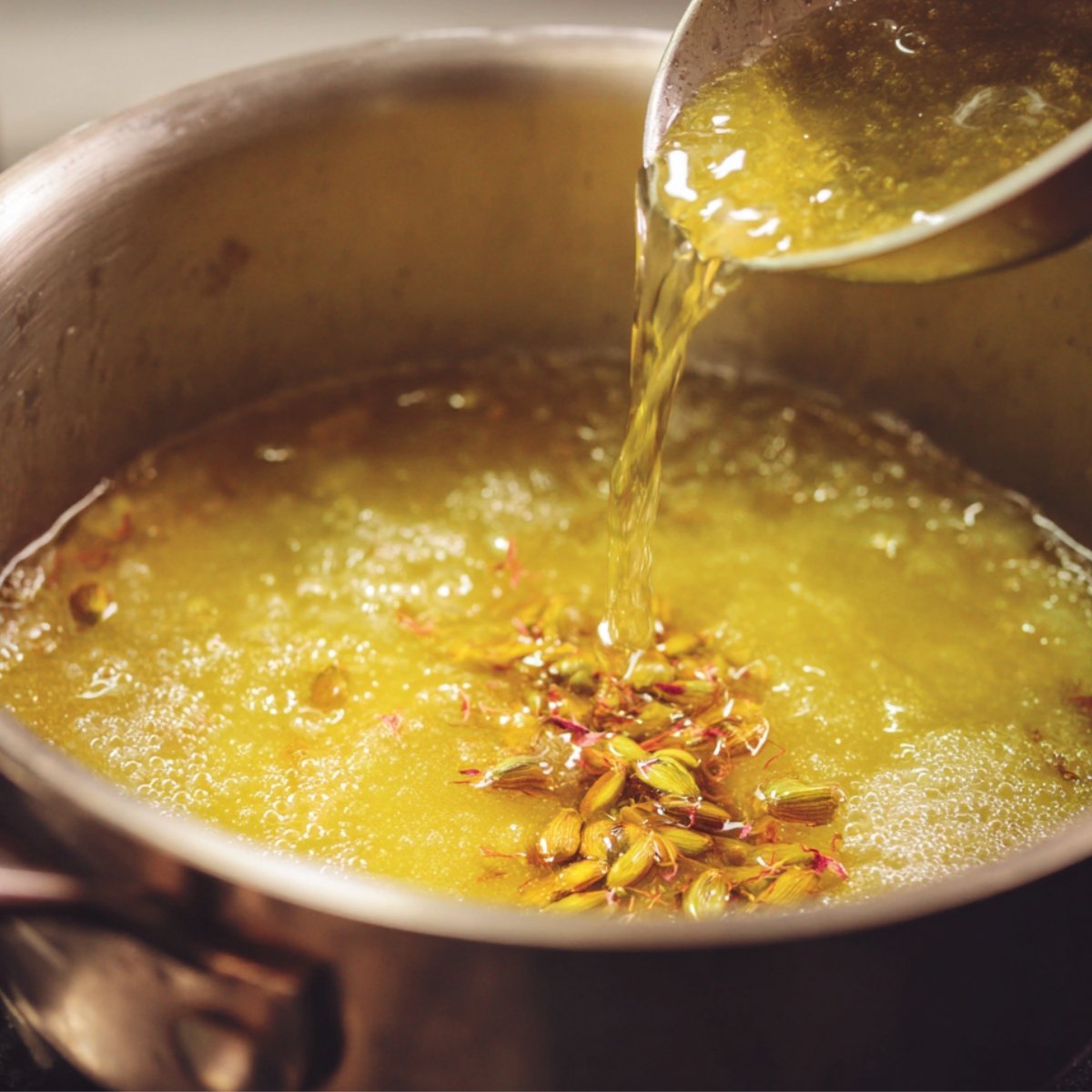 Sugar syrup with cardamom pods and saffron being boiled in a pot, while more syrup is poured in.