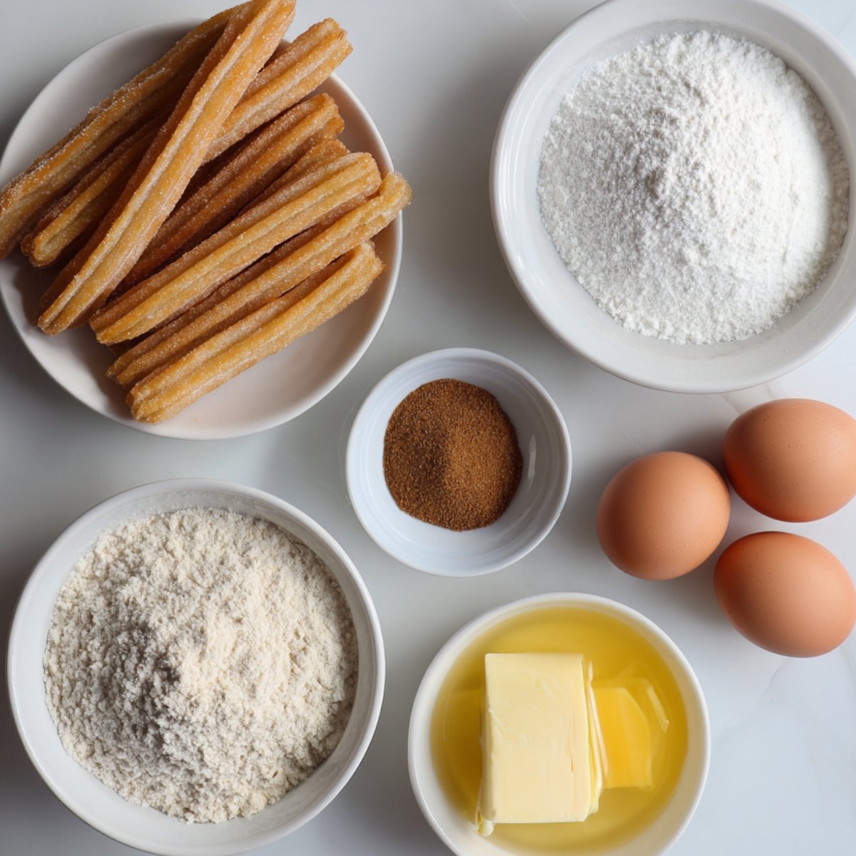 Baking ingredients for churros on a white surface: eggs, flour, butter, sugar, and cinnamon.