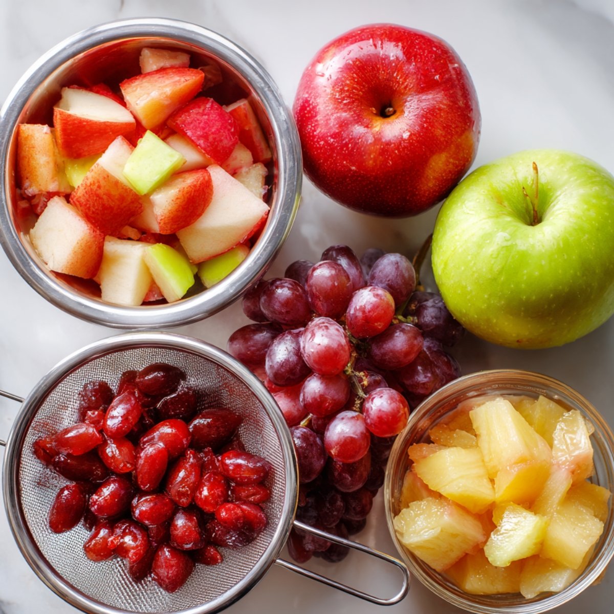 Apples, grapes, cranberries in a strainer, and pineapple chunks laid out for salad preparation.