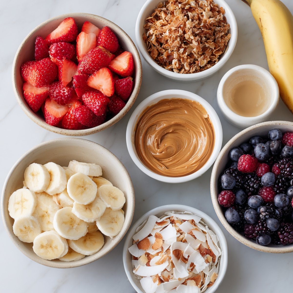 An assortment of toppings in bowls including sliced bananas, strawberries, mixed berries, granola, peanut butter, coconut flakes, and a banana, ready to be added to chia seed pudding.