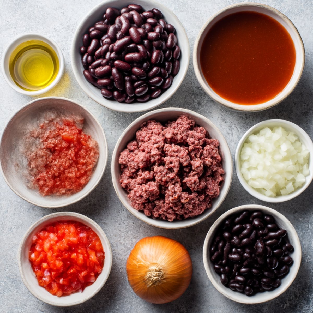 A set of bowls with black beans, tomatoes, ground beef, diced onions, and tomato sauce, along with olive oil, arranged on a clean surface for preparing chili.