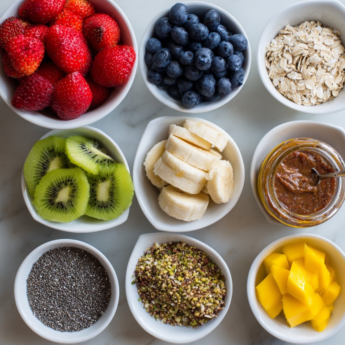 An assortment of toppings in small bowls: strawberries, blueberries, banana slices, kiwi, mango, chia seeds, oats, pistachios, and nut butter.