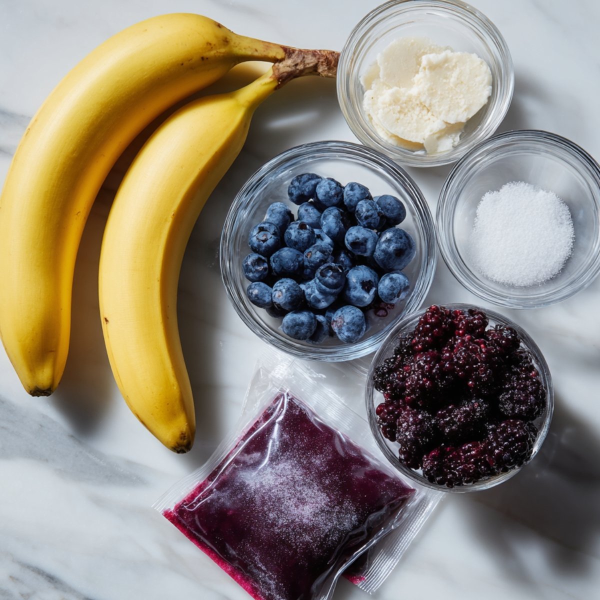 Ingredients for acai bowl on a marble surface: whole bananas, frozen acai packet, blueberries, blackberries, shredded coconut, and sugar.