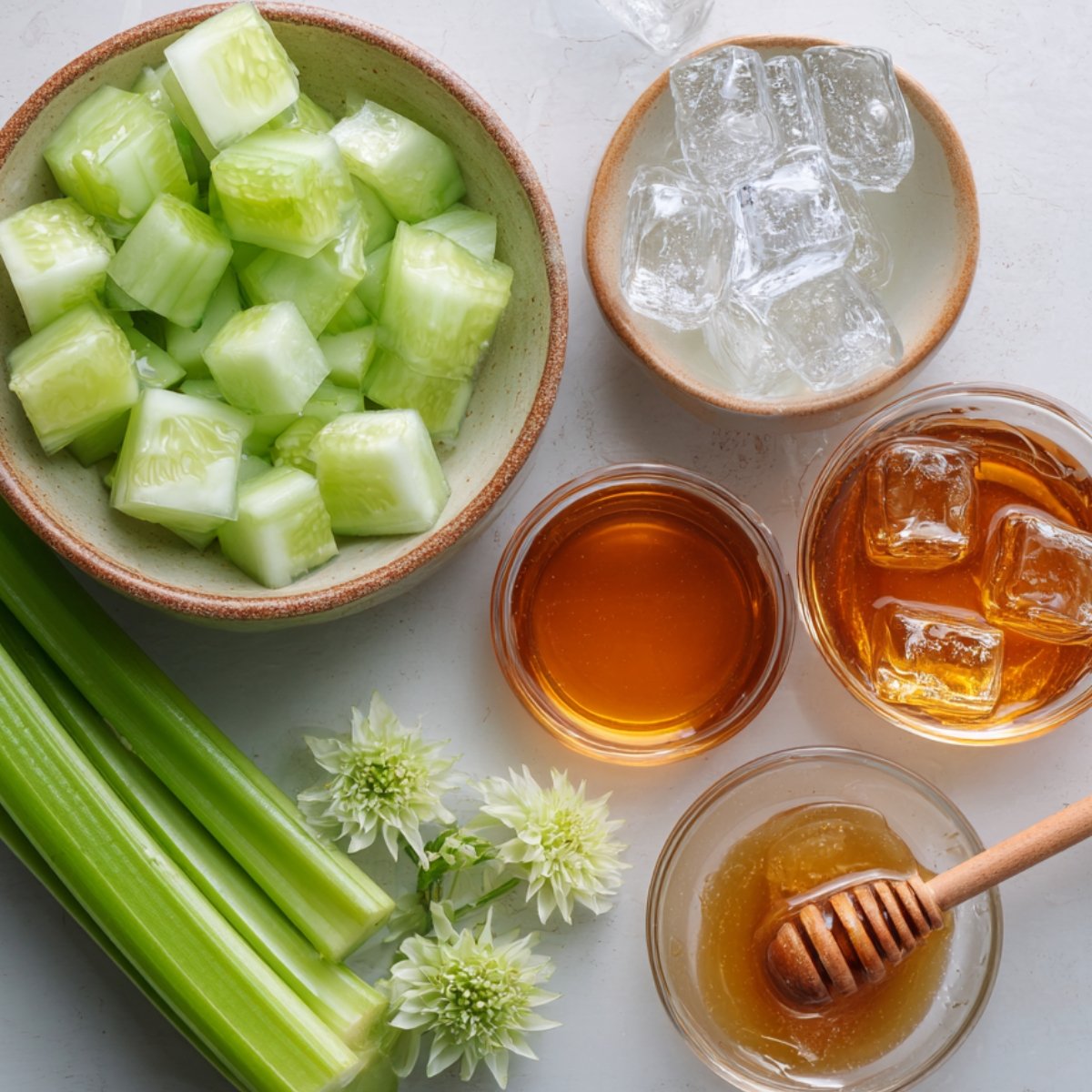Fresh cucumber chunks, celery stalks, bowls of honey, ice cubes, and decorative flowers arranged on a white surface.