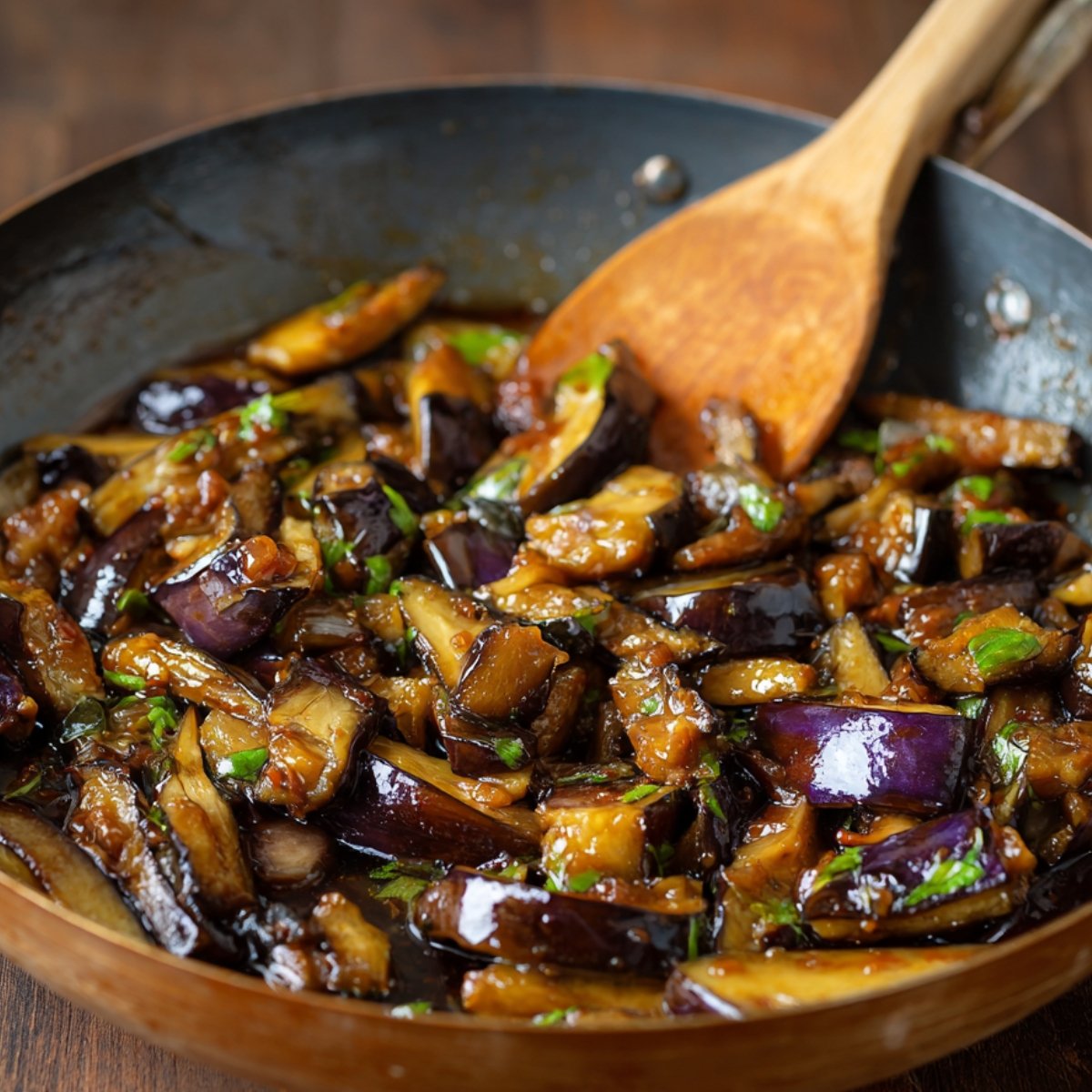 A wok filled with stir-fried Japanese eggplant coated in a savory sauce, with a wooden spoon mixing the dish.