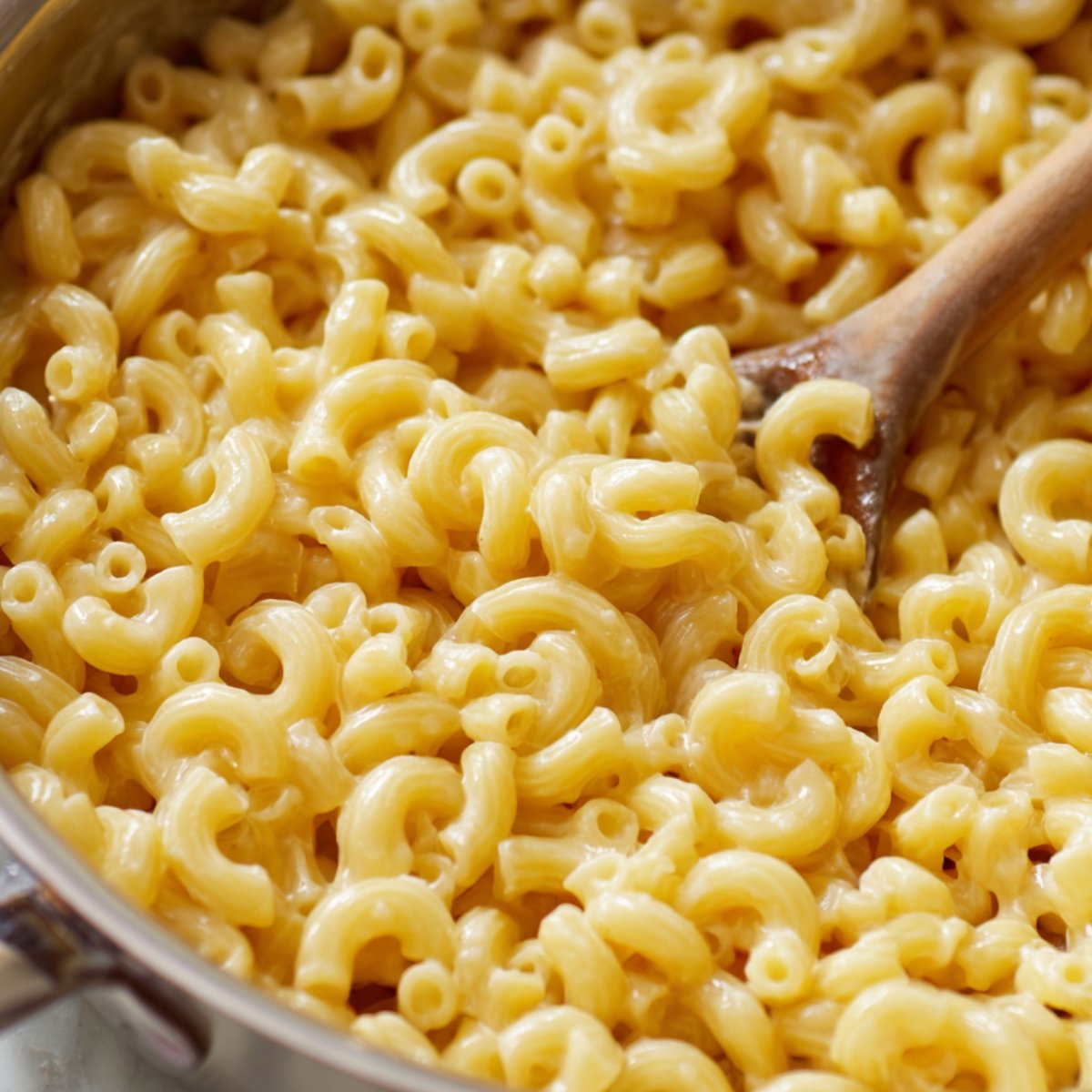 A pot of cooked elbow macaroni coated in creamy cheese sauce, stirred with a wooden spoon.
