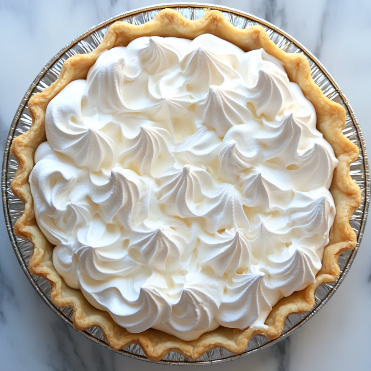 A lemon meringue pie topped with fluffy, glossy white meringue swirls that have not been toasted, in a foil pie tin on a marble surface.